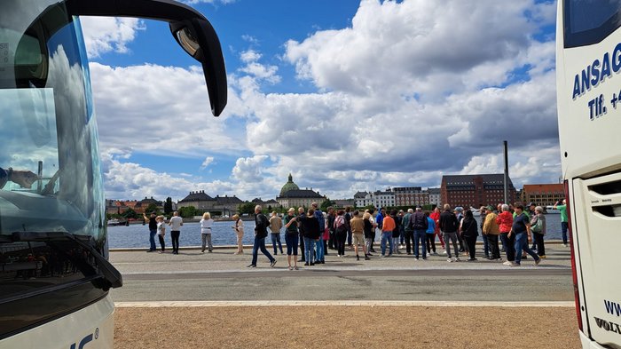 Reisefreudige Althandwerker/innen Menschen stehen am Wasserufer zwischen zwei Bussen mit Stadtkulisse im Hintergrund
