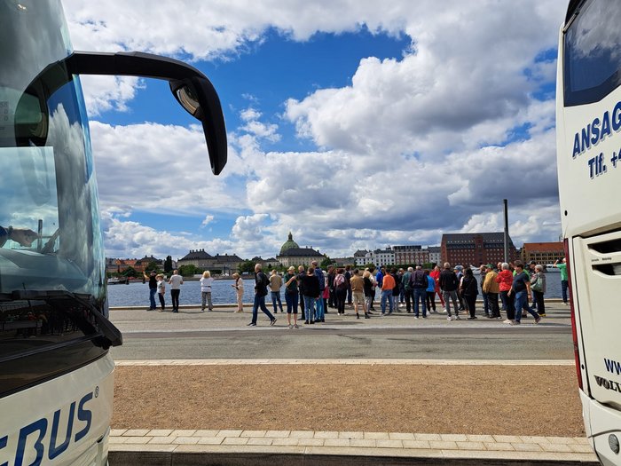 Reisefreudige Althandwerker/innen Menschen stehen am Wasserufer zwischen zwei Bussen mit Stadtkulisse im Hintergrund