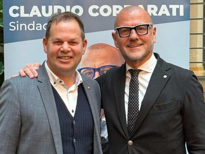 „Gemeindewahlen in Bozen: eine starke Stimme für das Handwerk“ Zwei Männer in Anzügen vor einem Wahlplakat von Claudio Corrarati