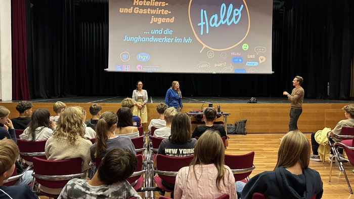 Jugendliche in Eppan entdecken Berufe im Handwerk und Gastgewerbe Jugendliche sitzen in einem Saal vor einer Bühne mit Präsentation und sprechen mit drei Erwachsenen