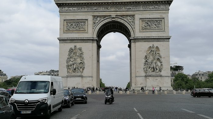 lvh-Seniorinnen und Senioren auf Frühlingsreise: Paris, mon amour! Ansicht des Arc de Triomphe mit parkenden Autos und Menschen bei bewölktem Himmel