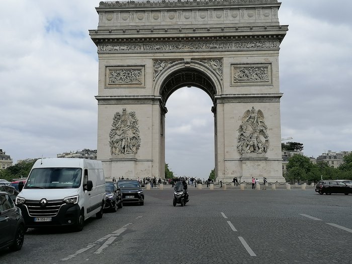 lvh-Seniorinnen und Senioren auf Frühlingsreise: Paris, mon amour! Ansicht des Arc de Triomphe mit parkenden Autos und Menschen bei bewölktem Himmel