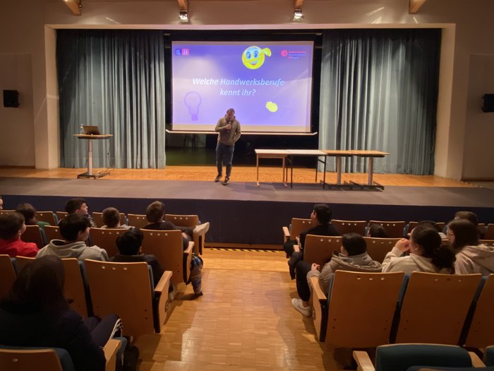 Mittelschulen Mals und Graun im Vinschgau: Handwerk & Gastgewerbe hautnah erleben. Eine Initiative der Junghandwerker/innen und der HGJ. Mann spricht vor Publikum in einem Vortrag über Handwerksberufe