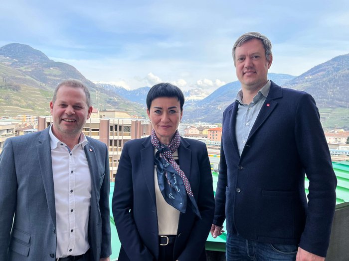 Cooperazione per l'edilizia sociale: lvh.apa incontra l'assessora provinciale Ulli Mair Tre persone in abbigliamento formale con paesaggio montano sullo sfondo