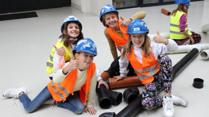 Vivere una giornata in cantiere Bambini con caschi e giubbotti di sicurezza posano con tubi facendo il segno del pollice in su