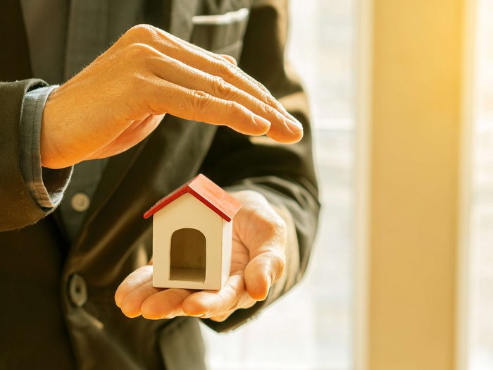 Katastrophenversicherungen: lvh fordert weiteren Aufschub Person schützt mit Hand ein kleines Hausmodell im Anzug