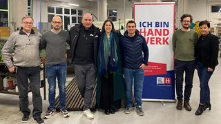 News Sieben Personen stehen in einer Werkstatt vor einem Ich bin Handwerk Banner