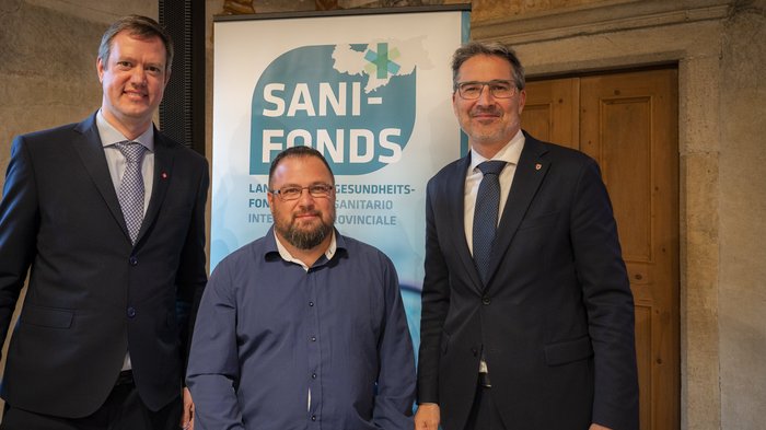 10 Jahre Sani-Fonds Drei Männer stehen vor einem Sani-Fonds Banner in formeller Kleidung