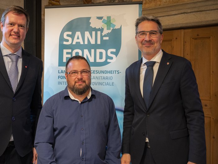 10 Jahre Sani-Fonds Drei Männer stehen vor einem Sani-Fonds Banner in formeller Kleidung