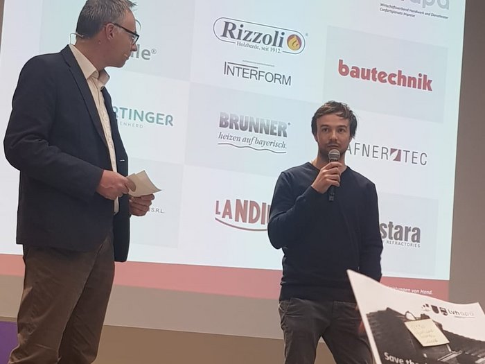 Aktivitäten der Hafner/innen im lvh Zwei Männer sprechen vor Sponsorentafel bei einer Veranstaltung