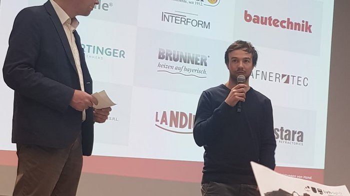 Aktivitäten der Hafner/innen im lvh Zwei Männer sprechen vor Sponsorentafel bei einer Veranstaltung