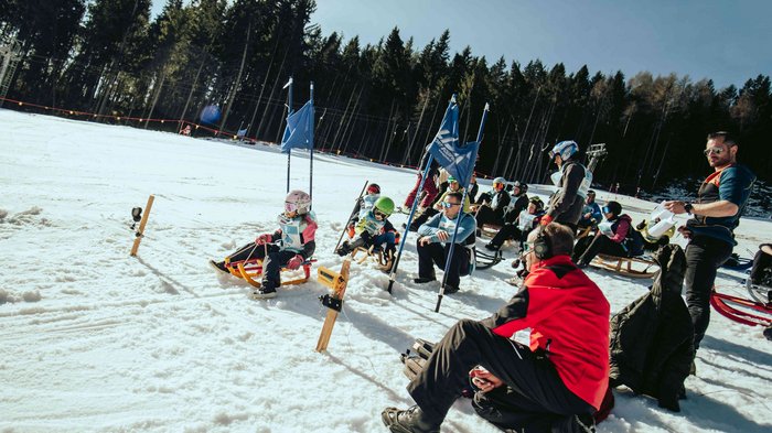 lvh Wintersportfest in Feldthurns Kinder und Erwachsene beim Rodelrennen auf verschneiter Piste mit Wald im Hintergrund