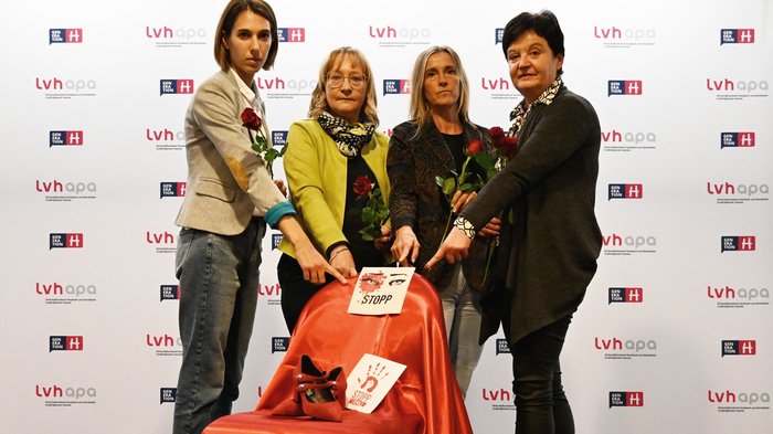 lvh setzt Zeichen gegen Gewalt an Frauen: ein „Besetzter Platz“ Vier Frauen mit Rosen zeigen auf rot bedeckten Stuhl mit Stoppschildern