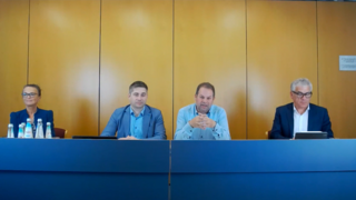 Webinare Vier Personen sitzen hinter einem Konferenztisch vor einer Holzpaneelwand