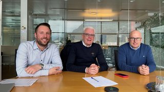 News Drei Männer sitzen an einem Konferenztisch mit Dokumenten und Geräten