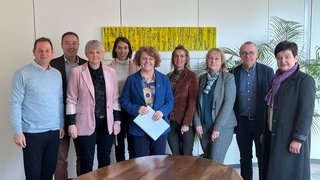 News Gruppe von neun Personen steht in einem Besprechungsraum vor einem ovalen Holztisch