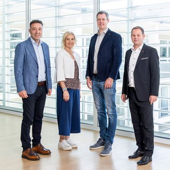 lvh-Geschichte Vier Personen in Businesskleidung stehen in einem hellen Büro vor großen Fenstern