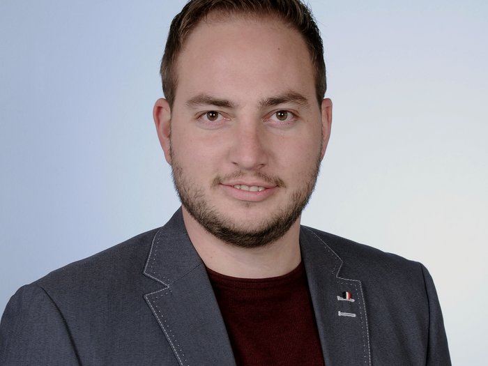 Tiefbauer beklagen untragbare Treibstoffpreise Porträt eines Mannes mit dunkelblauem Sakko und rotem Pullover vor hellblauem Hintergrund