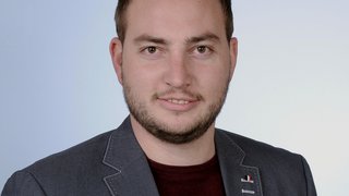 News Porträt eines Mannes mit dunkelblauem Sakko und rotem Pullover vor hellblauem Hintergrund