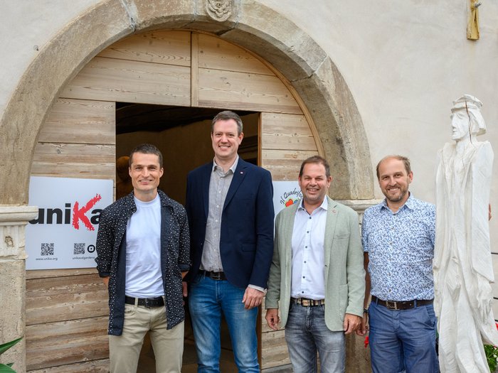 UNIKA in Livo: „Alt(r)e Visioni 4 – Dalla montagna all’arte“ 25 Künstler:innen stellen im Palazzo Aliprandini Laifenthurn aus Vier Männer stehen vor einem großen Holztor mit Steinrahmen und weißer Holzskulptur