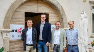 News Vier Männer stehen vor einem großen Holztor mit Steinrahmen und weißer Holzskulptur