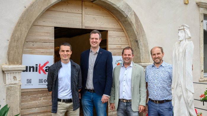 UNIKA a Livo: “Alt(r)e Visioni 4 – Dalla montagna all'arte” 25 artisti e artiste espongono a Palazzo Aliprandini Laifenthurn Quattro uomini in piedi davanti a un grande portone di legno con cornice in pietra e scultura in legno bianca