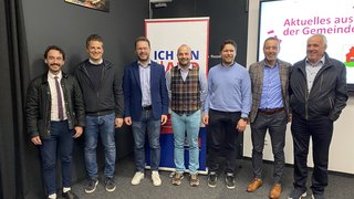 News Sieben Männer stehen nebeneinander vor Werbebannern und lächeln in die Kamera