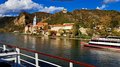 Auf Reisen: Althandwerker auf Flusskreuzfahrt Flusskreuzfahrtschiff vor historischer Stadt und Hügeln unter blauem Himmel