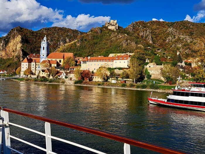 Auf Reisen: Althandwerker auf Flusskreuzfahrt Flusskreuzfahrtschiff vor historischer Stadt und Hügeln unter blauem Himmel