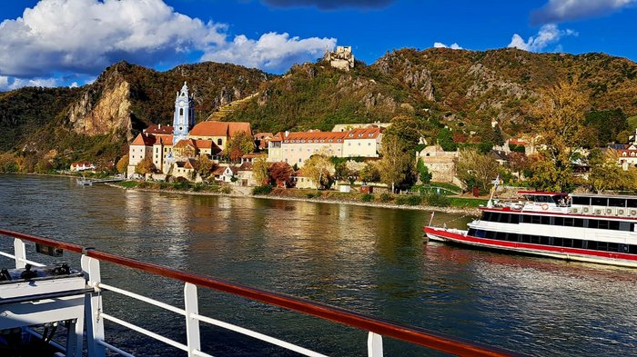 Auf Reisen: Althandwerker auf Flusskreuzfahrt Flusskreuzfahrtschiff vor historischer Stadt und Hügeln unter blauem Himmel