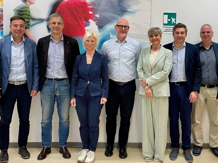 Incontriamoci a scuola! Nove persone in piedi in una stanza luminosa davanti a una parete con arte e uscita di emergenza.