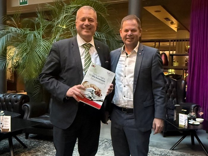 Pflichtversicherung gegen Naturkatastrophen – lvh erfreut über Aufschub Zwei Männer im Anzug stehen in einem Hotel-Lobby und halten ein Buch.