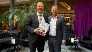 News Zwei Männer im Anzug stehen in einem Hotel-Lobby und halten ein Buch.