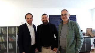 News Drei Männer stehen zusammen in einem Raum und lächeln in die Kamera