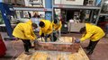 Campagna informazione carriere Nova Ponente e Nova Levante Quattro persone con giacche gialle costruiscono un muro di mattoni in una officina