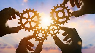 News Hände halten Zahnräder vor dem Sonnenuntergang, Symbol für Zusammenarbeit