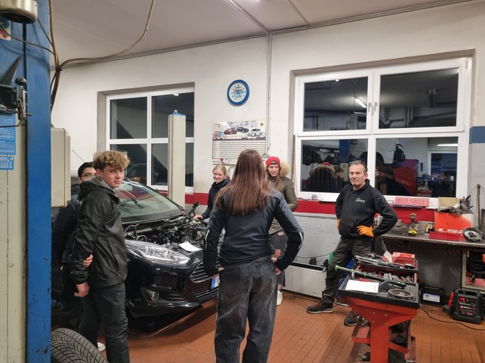 Erster „Open Day im Handwerk“ begeistert Jugendliche und Eltern Gruppe junger Leute in Autowerkstatt beim Gespräch mit Mechaniker vor Auto mit offener Motorhaube