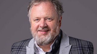 News Porträt eines lächelnden Mannes mit Bart und kariertem Sakko vor grauem Hintergrund