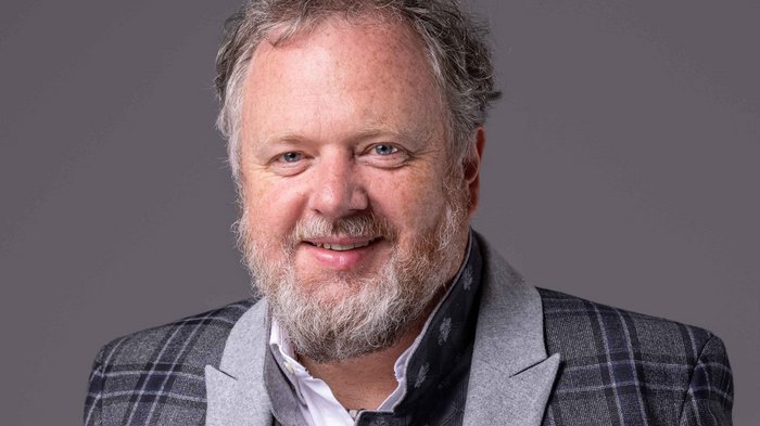 Für mehr Sicherheit am Arbeitsplatz/Lanz: „Mehr beraten und weniger strafen!“ Porträt eines lächelnden Mannes mit Bart und kariertem Sakko vor grauem Hintergrund