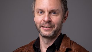 News Porträt eines lächelnden Mannes mit brauner Lederjacke vor grauem Hintergrund