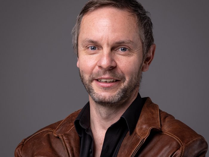 Großes Interesse für Öffentliche Aufträge im Pustertal Porträt eines lächelnden Mannes mit brauner Lederjacke vor grauem Hintergrund