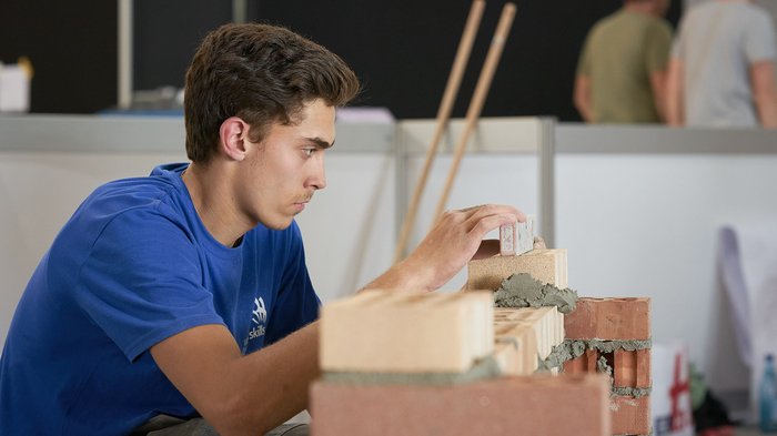 Handwerk live: Junge Talente zeigen, was sie draufhaben Junger Bauarbeiter mauert konzentriert eine Wand aus Ziegelsteinen im Innenbereich