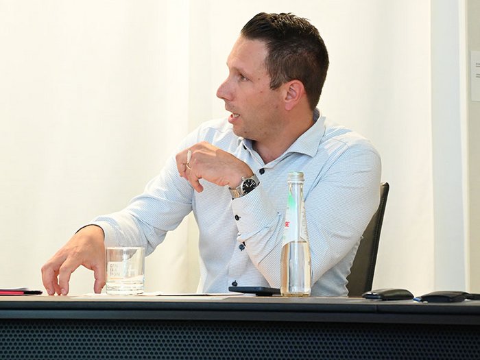 Wohnen muss leistbar sein! Herausforderungen und Lösungsansätze in Südtirol Mann im Hemd sitzt am Tisch und spricht, mit Glas und Flasche davor