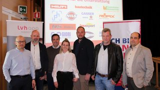 News Sieben Personen posieren vor einer Wand mit Sponsorlogos bei einer Veranstaltung