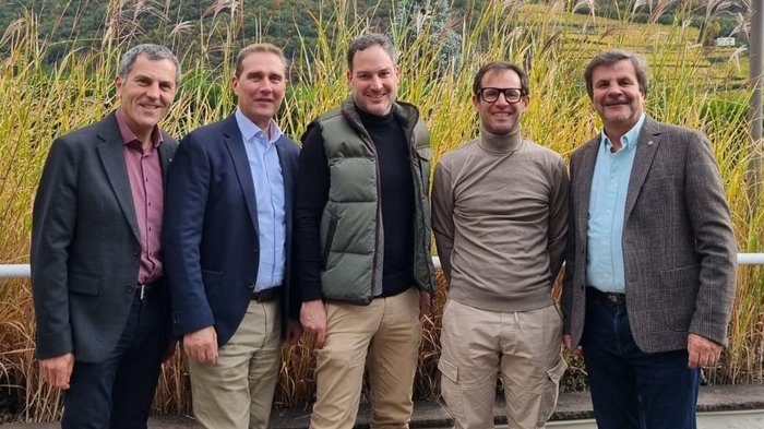 Insieme per un futuro sicuro dal punto di vista energetico: incontro trilaterale dei maestri spazzacamini in Alto Adige Cinque uomini sorridenti in piedi davanti a erba alta all'aperto