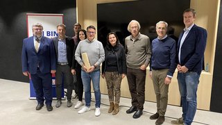 News Gruppe von neun Personen steht vor Wand mit Fernsehbildschirm und Banner