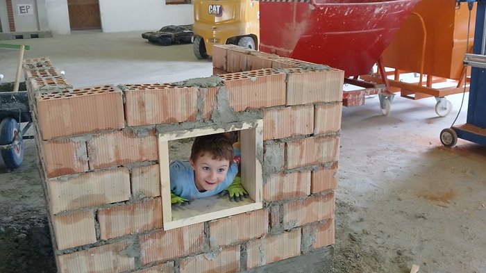 Kids-Academy 2024: buntes Handwerk in Bruneck Kind schaut durch kleines Fenster in halbfertiger Ziegelmauer in Baustellenhalle