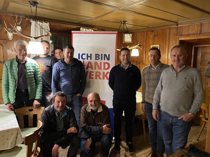 Terenten: Handwerk und Gemeindeentwicklung im Fokus Neun Männer posieren vor einem Ich-bin-Land-Handwerk-Banner in einem Holzraum.