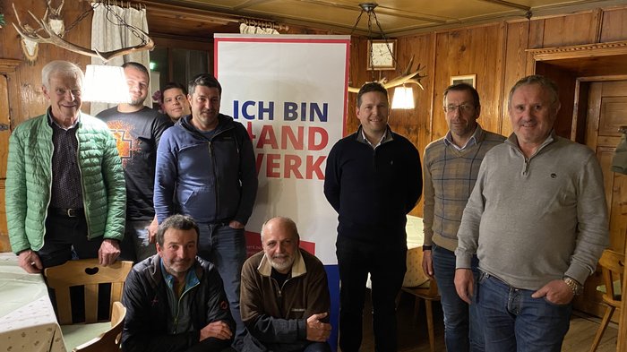 Terenten: Handwerk und Gemeindeentwicklung im Fokus Neun Männer posieren vor einem Ich-bin-Land-Handwerk-Banner in einem Holzraum.
