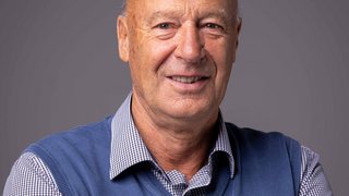 News Porträt eines lächelnden älteren Mannes mit blauem Pullover und kariertem Hemd vor grauem Hintergrund